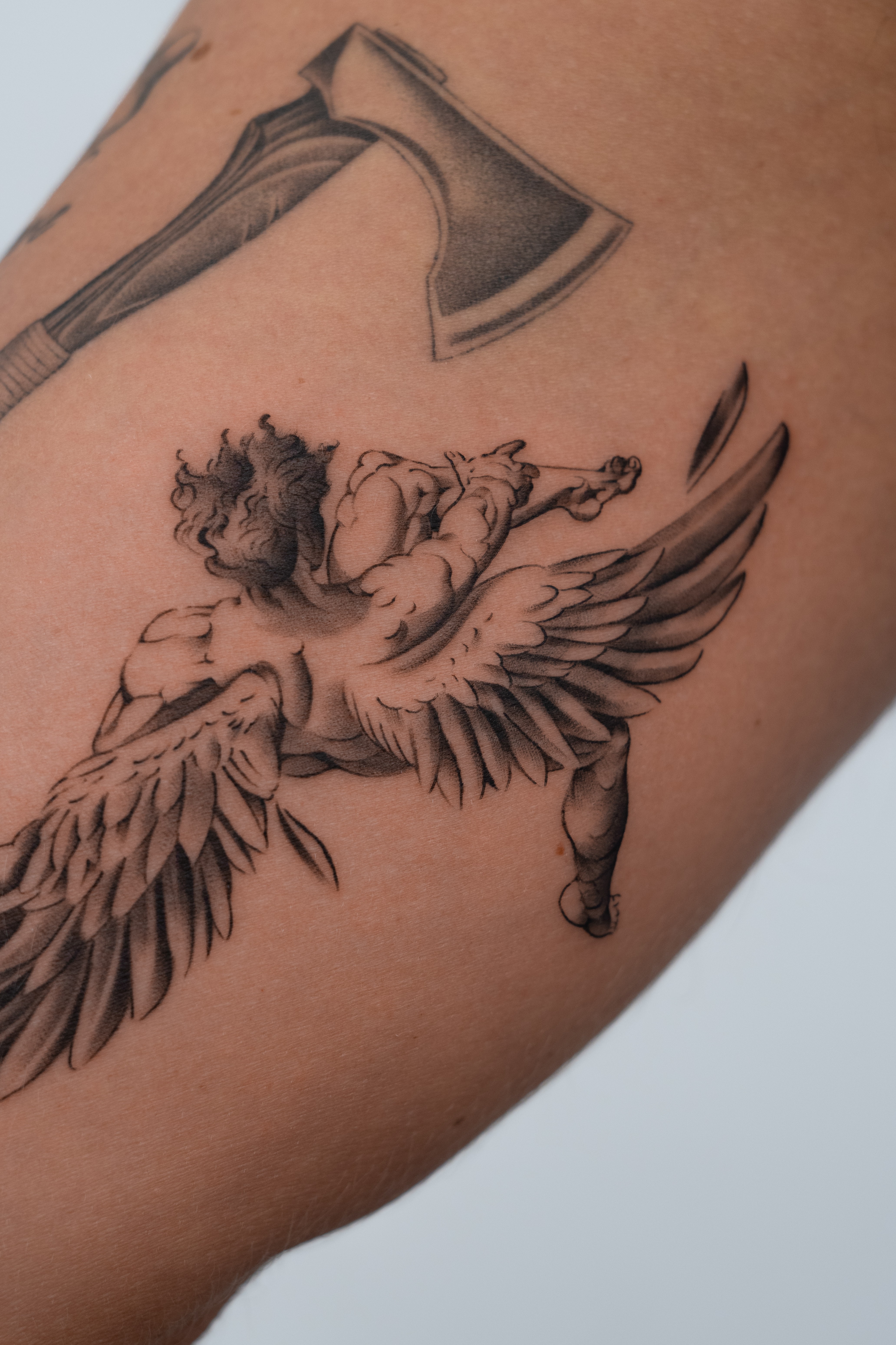 Fine-line cherub and axe tattoo on upper arm
